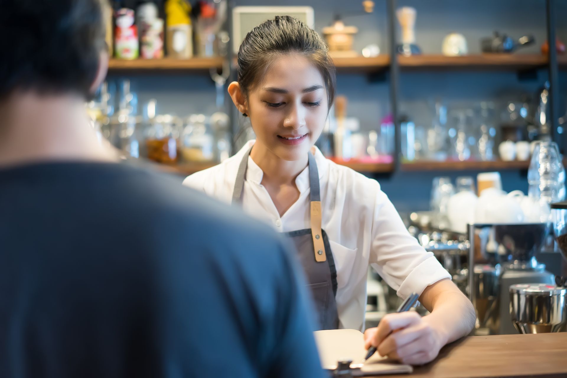 Barista in a white shirt preparing a drink behind a café counter while a customer sits in the foreground