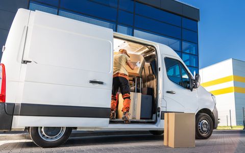 White delivery van with open side door in front of a blue building, boxes on the ground