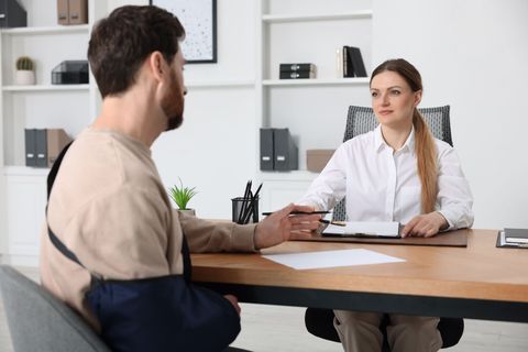 Two people in a medical office talking across a desk with papers and a clipboard