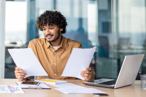 Man in a tan shirt reviewing papers at a desk with a laptop in a bright office