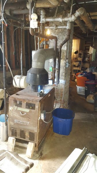 Old furnace in a basement setting with various pipes and clutter around it.