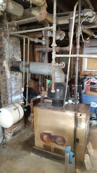 Old, gold-colored furnace in a basement setting with various pipes and fixtures.