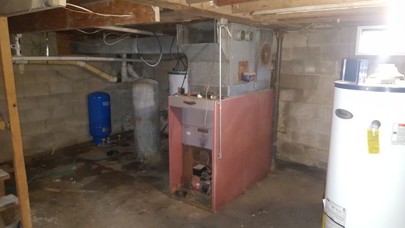 A basement with water heater, furnace, and water tank. Concrete walls and floor, utility pipes overhead.