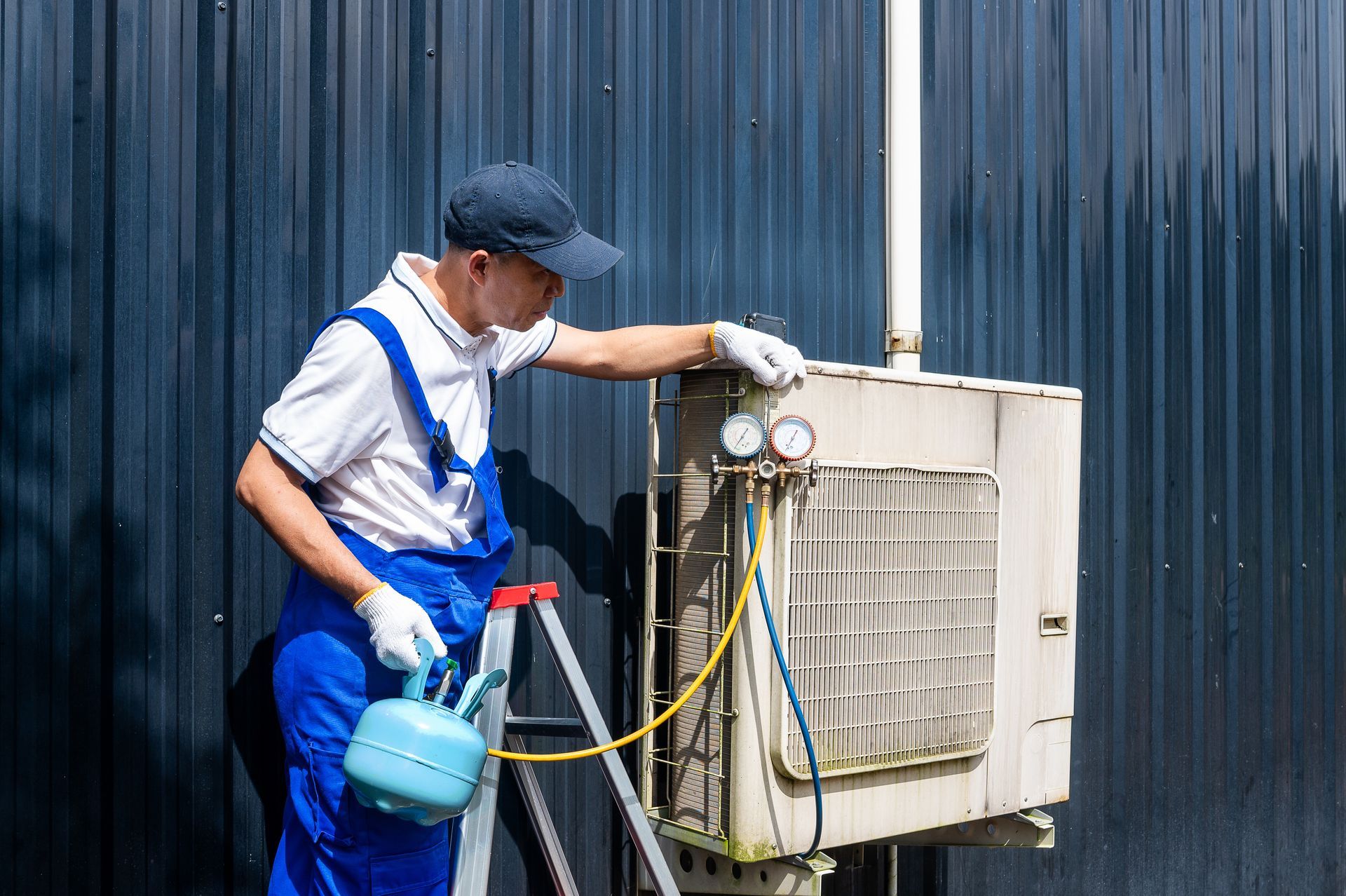 HVAC technician servicing outdoor unit. He’s wearing blue overalls and a cap. He's holding a blue canister and looking at gauges.