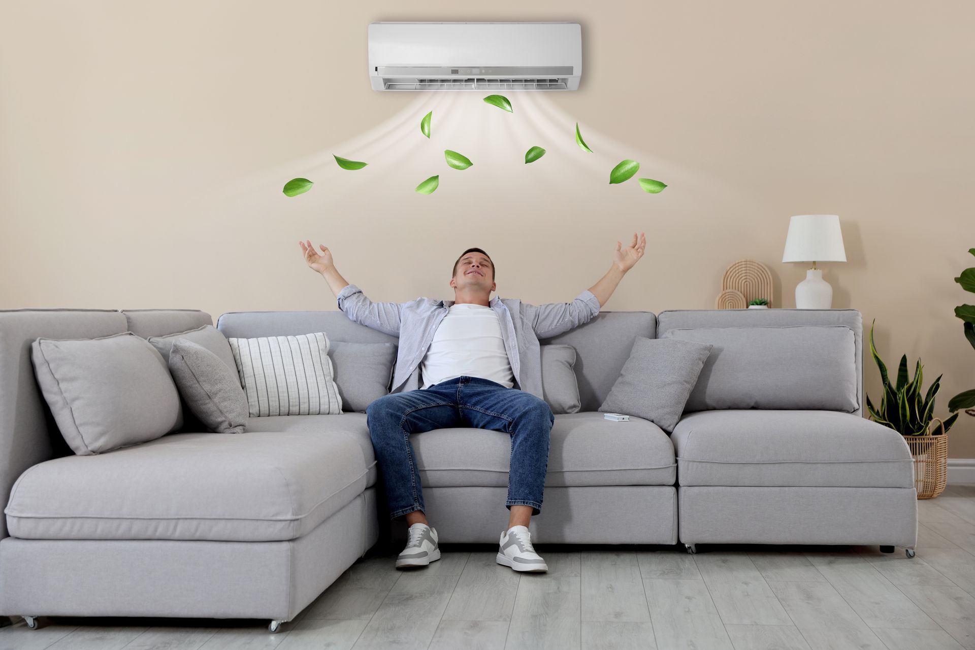 Man sitting on a sofa with arms raised, enjoying air from a wall-mounted air conditioner; green leaves float down.