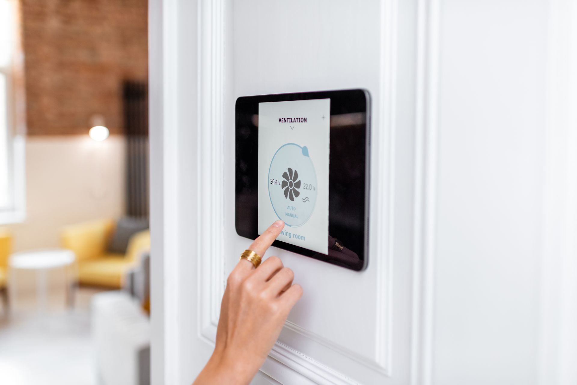 A person's hand touches a tablet mounted on a white wall, controlling a smart home feature.