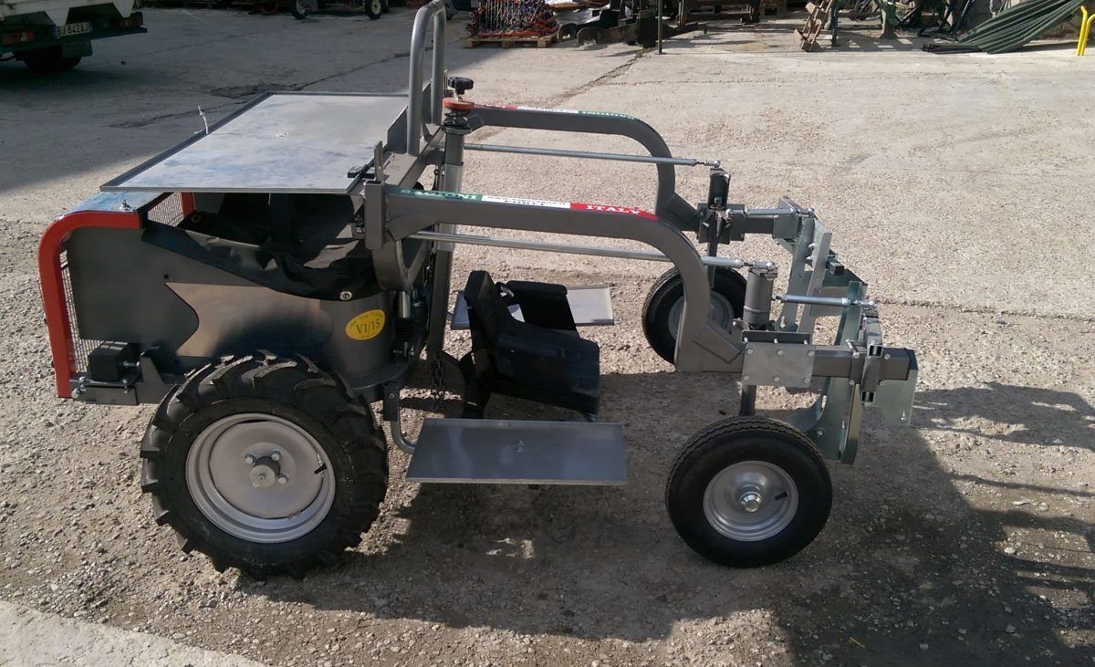 Vista laterale di una mietitrice o di un veicolo di trasporto agricolo grigio, a basso profilo e alimentato a batteria, su una superficie ghiaiosa.