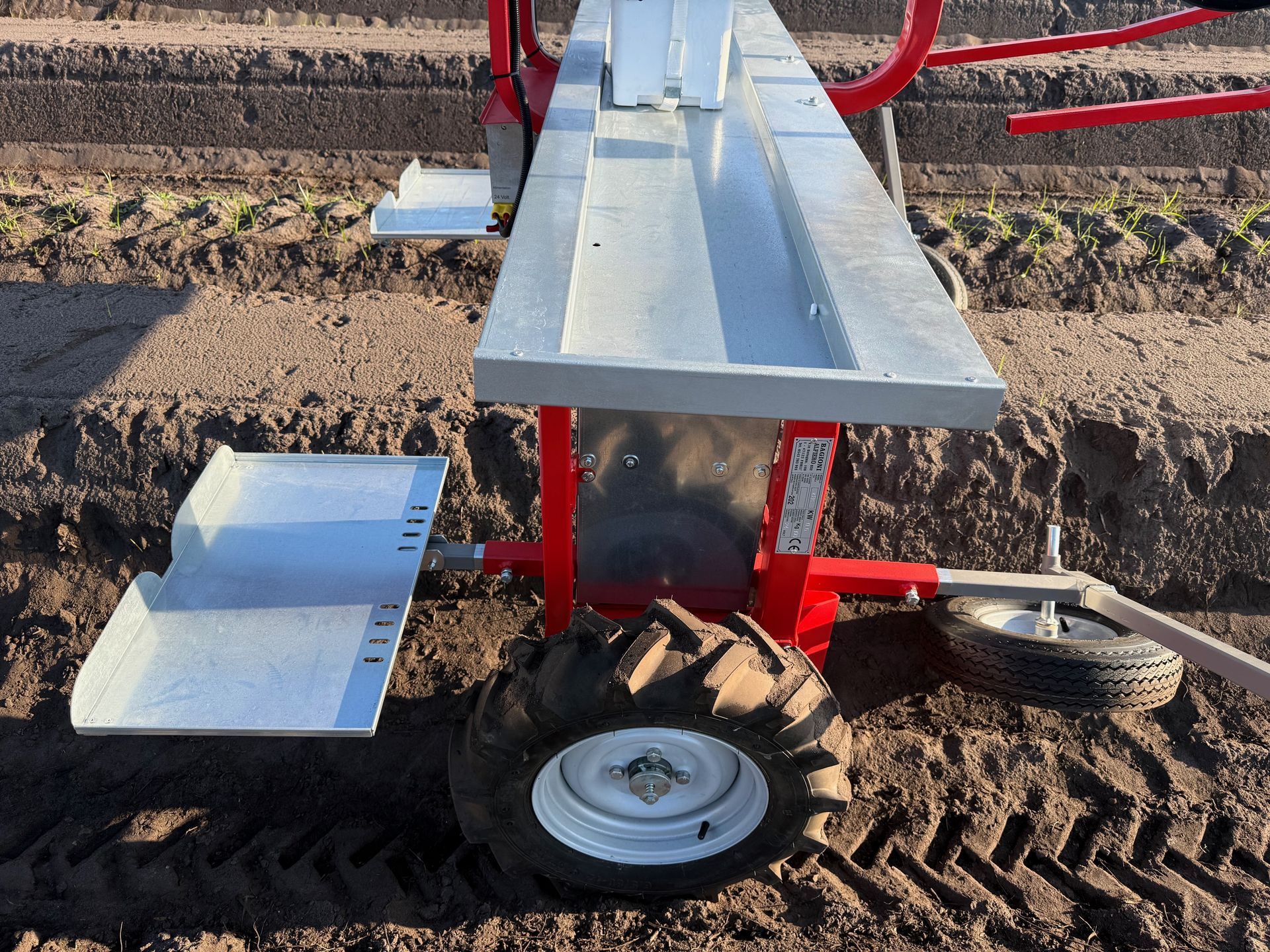 Una seminatrice agricola rossa e grigia si erge in un campo sterrato, con un piccolo pneumatico nero e una piattaforma metallica a vassoio.