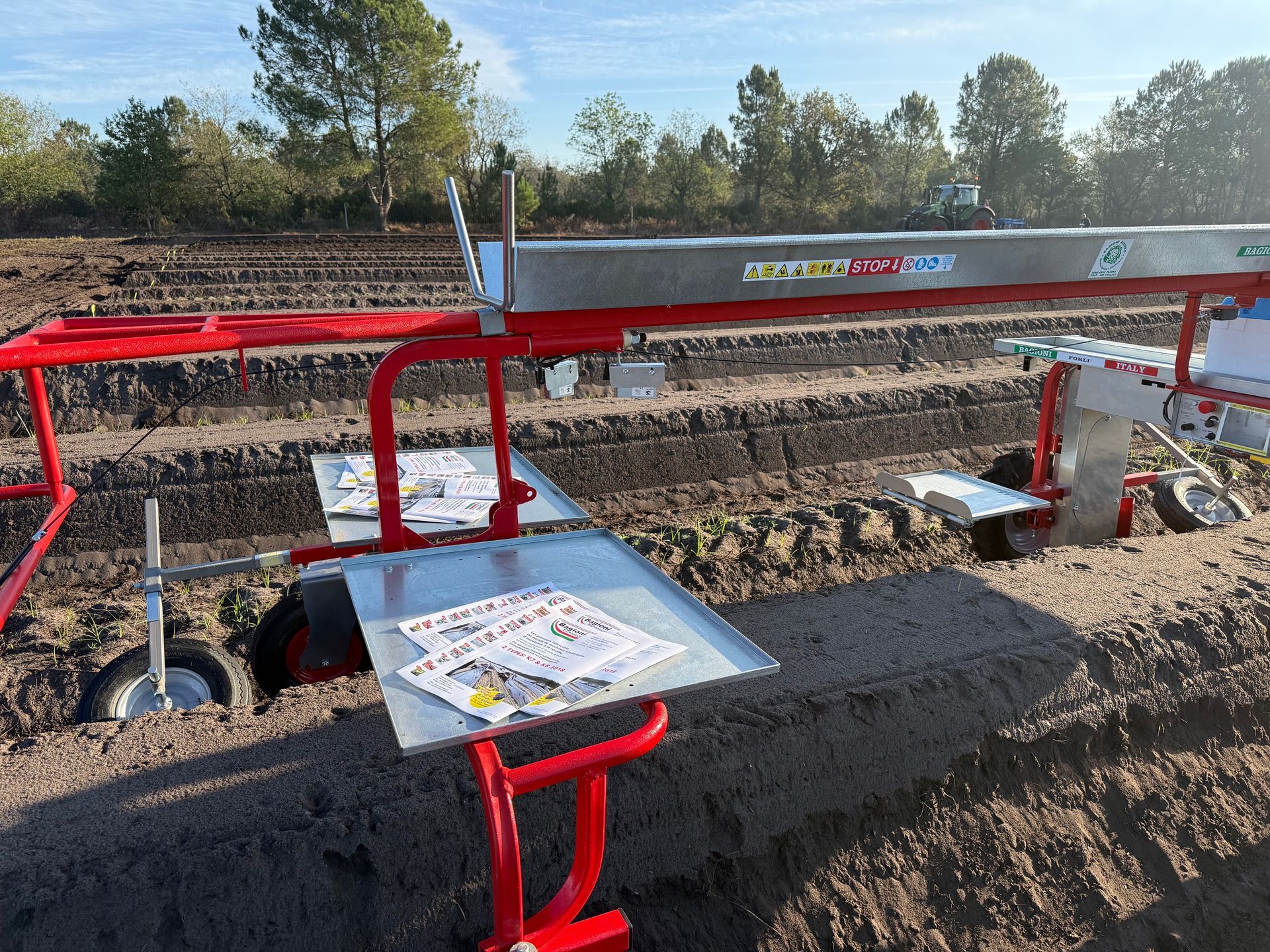 In un campo, sotto un cielo azzurro limpido, un telaio rosso per attrezzi agricoli con vassoi metallici poggia su file di terreno arato.