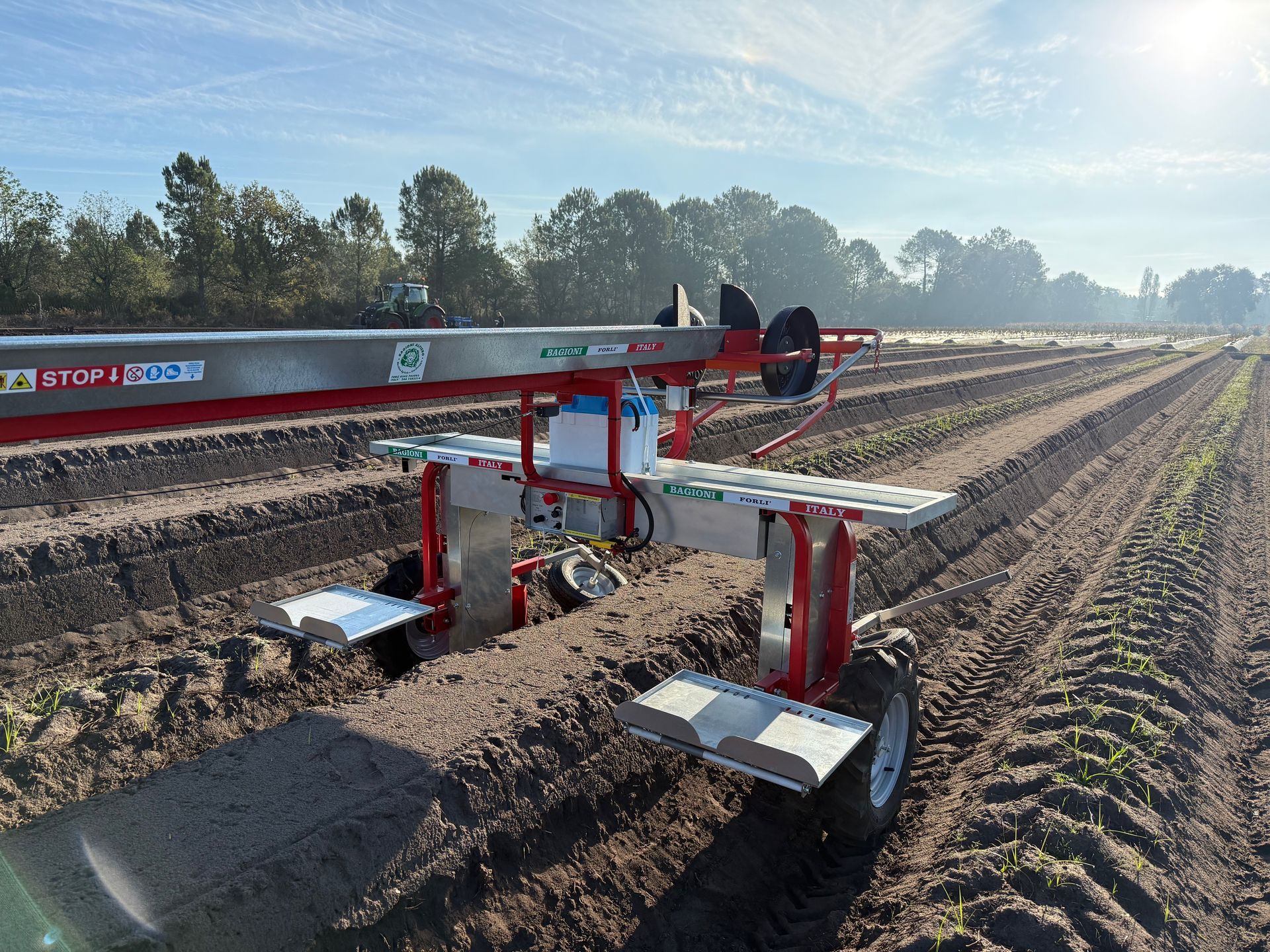 Una mietitrice agricola con telaio rosso e argento si muove attraverso un campo arato sotto un luminoso sole mattutino.