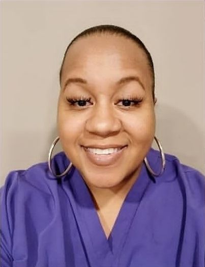 Woman with shaved head wearing a purple top, smiling, and wearing silver hoop earrings.