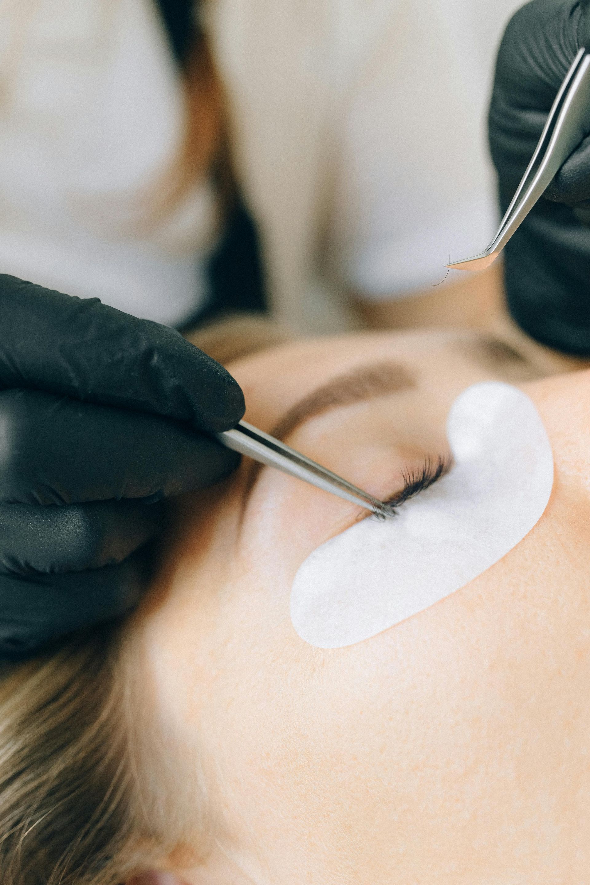 A person receiving eyelash extensions. A technician uses tweezers with gloved hands in close up.