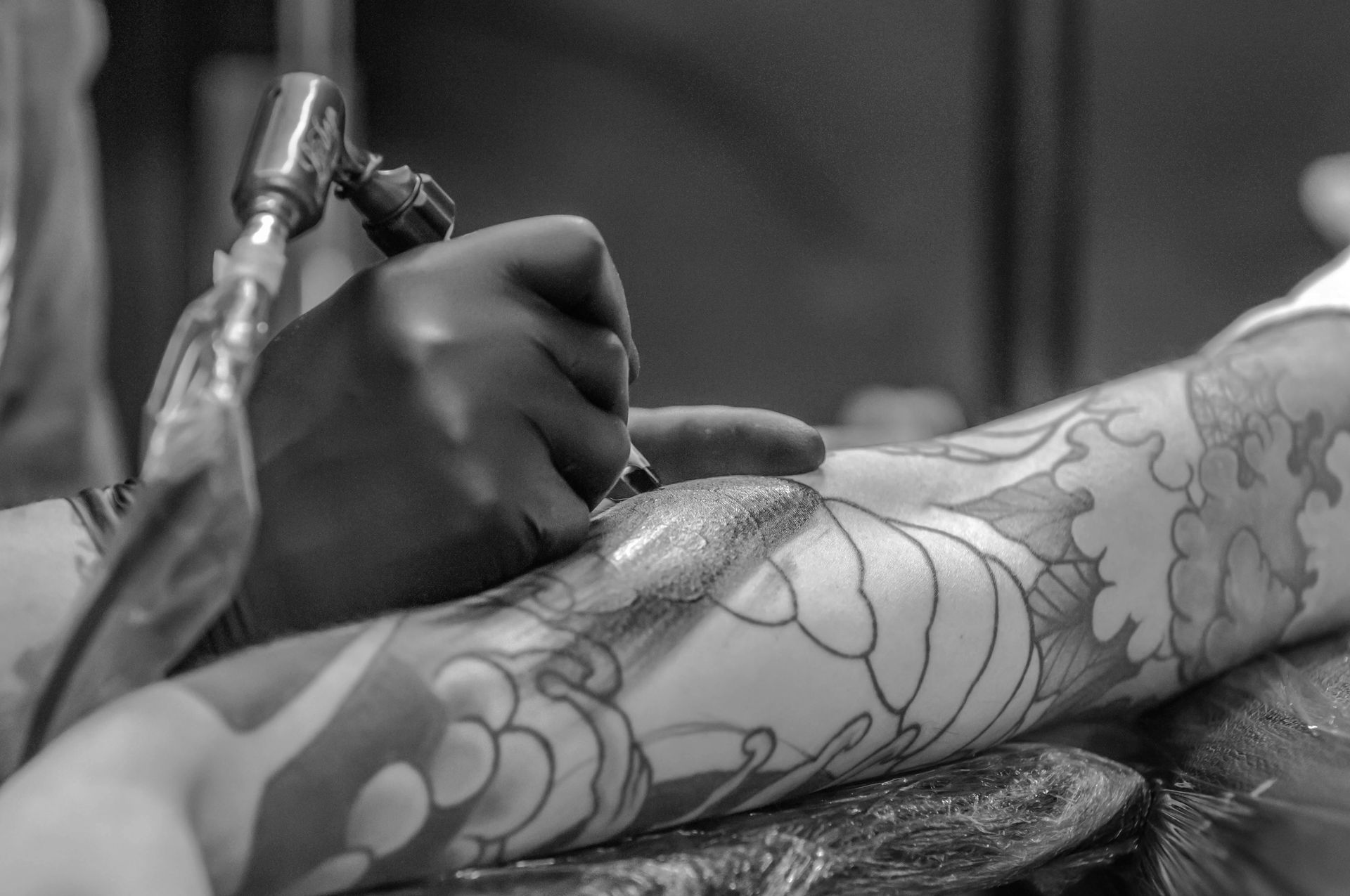 Tattoo artist in black gloves applying ink to an arm with a floral design, black and white.