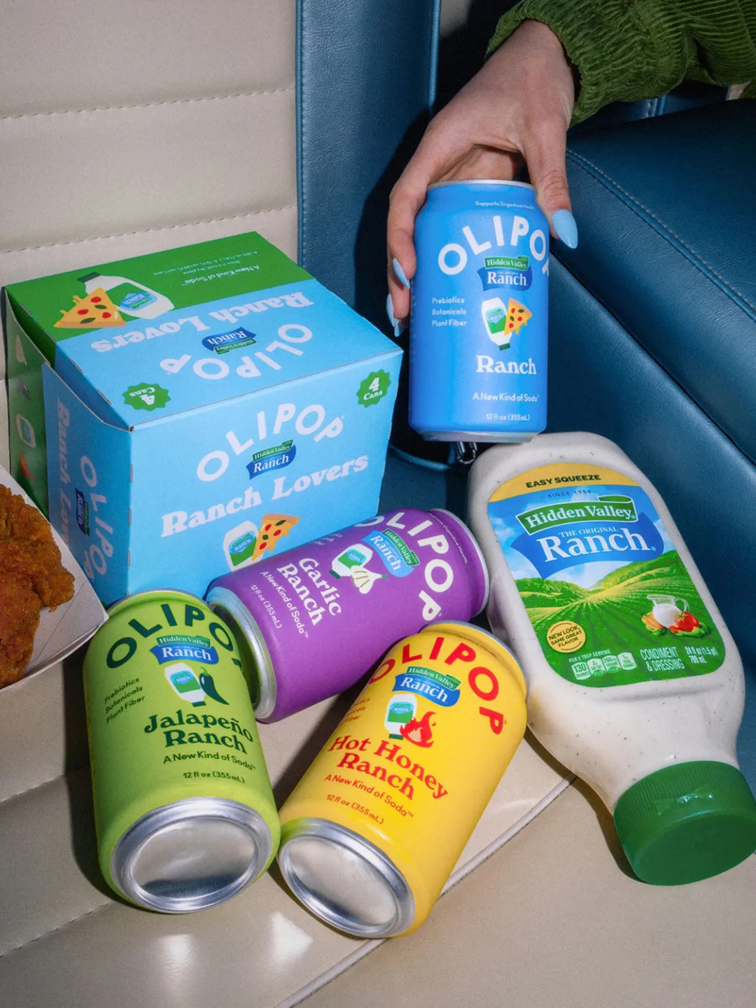 A hand holds a blue Olipop Ranch can next to a variety pack, three other flavored cans, and a bottle of ranch dressing.