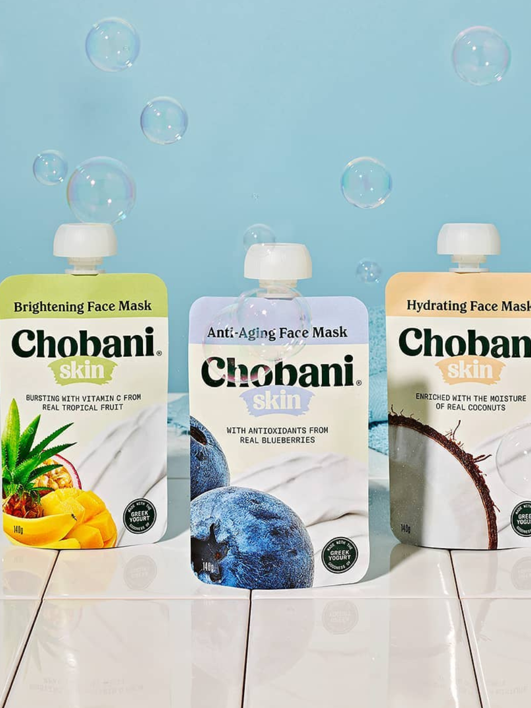 Three Chobani Skin face mask pouches in pineapple, blueberry, and coconut flavors set against a light blue, bubbly background.