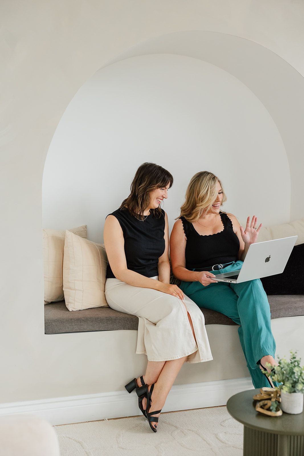 Two women smiling at a laptop. One waves, seated in a curved alcove. 