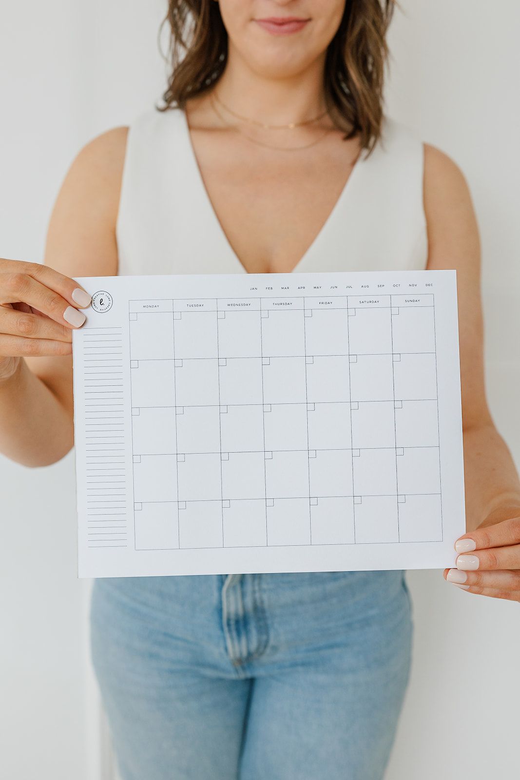 Woman holding a blank calendar against a white background; wearing a white top and jeans.