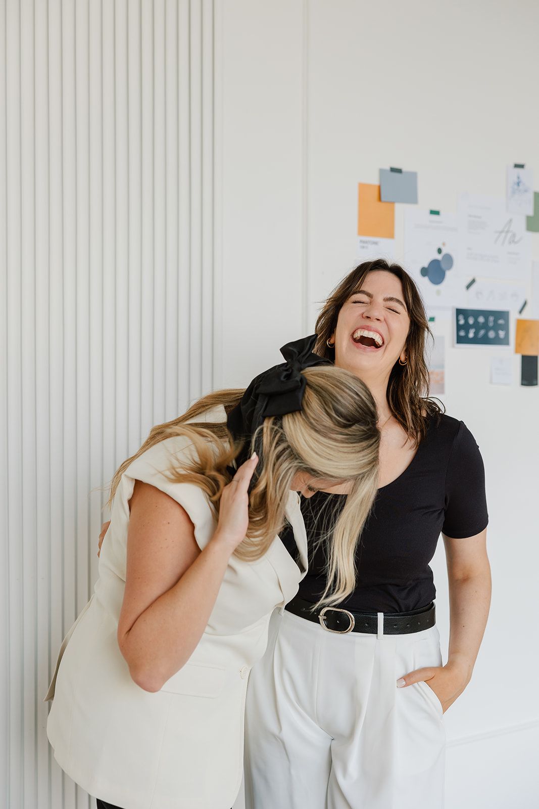 Two women laughing, one leaning, other's hand in pocket, white wall and mood board in background.