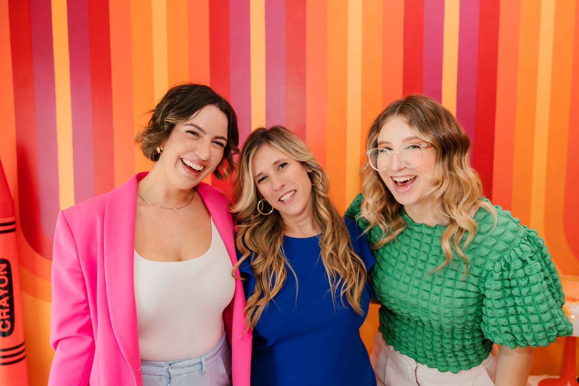 Three smiling women stand in front of a colorful striped backdrop.