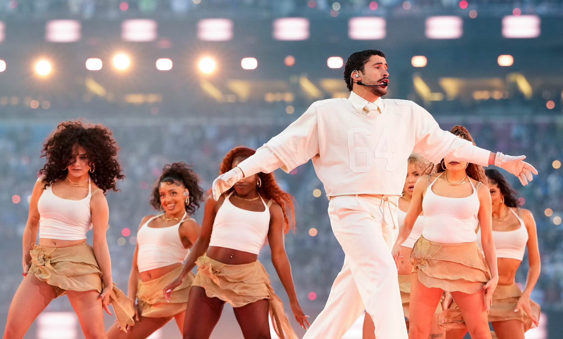 Singer in white sweater and pants performs on stage with backup dancers in a stadium.