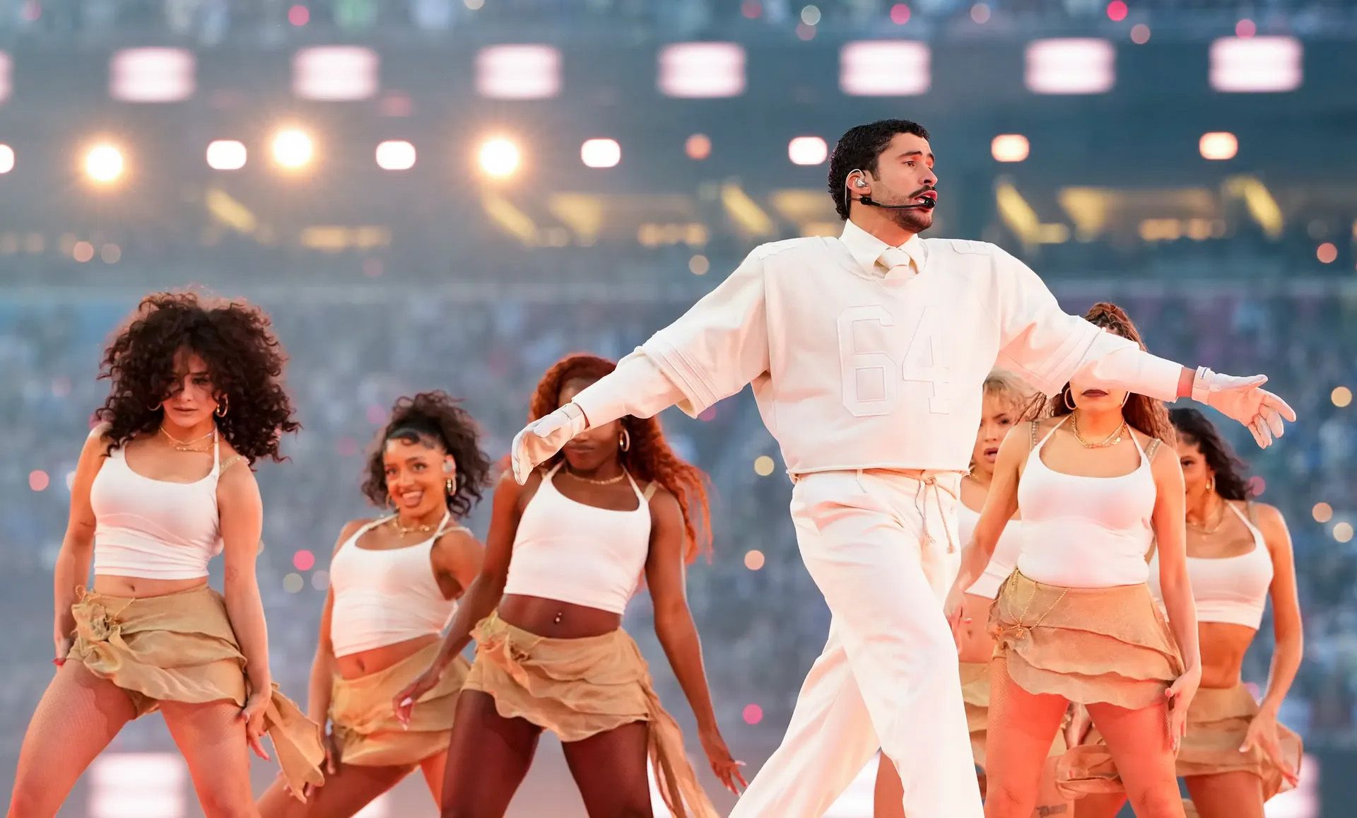 Singer in white sweater and pants performs on stage with backup dancers in a stadium.