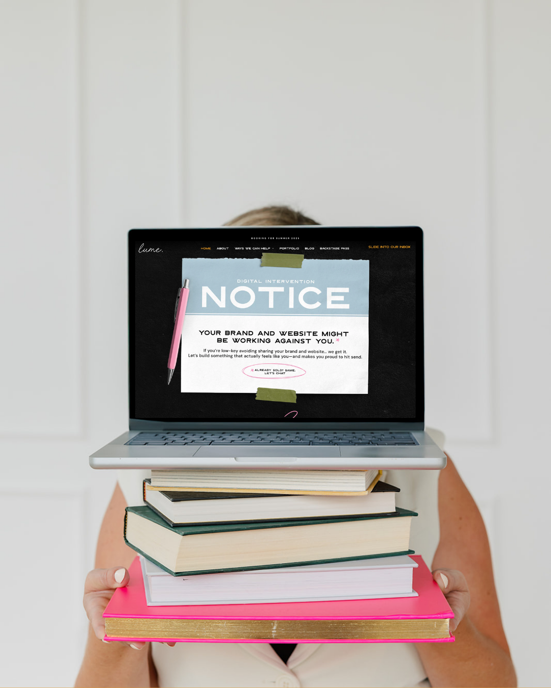A woman is holding a stack of books and a laptop that displays a custom website redesign
