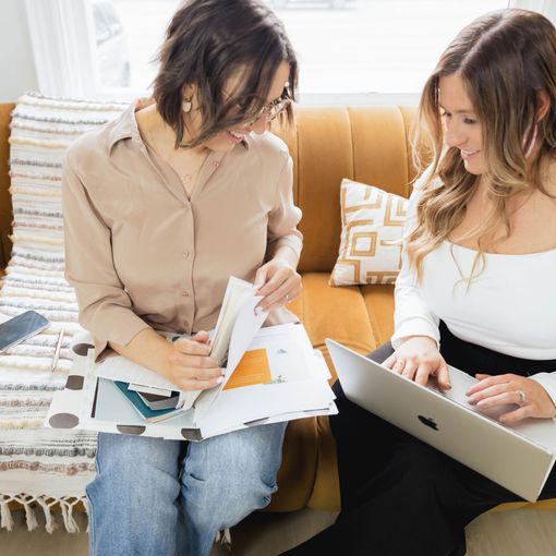 Two women are sitting on a couch looking at papers and a laptop