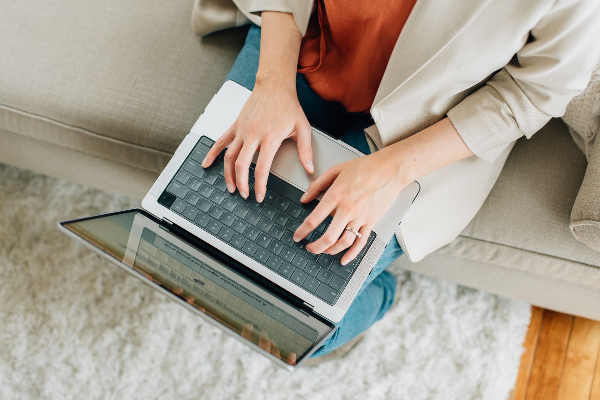 Person sitting on a couch with a laptop, typing. Wearing jeans, orange top, and tan blazer.