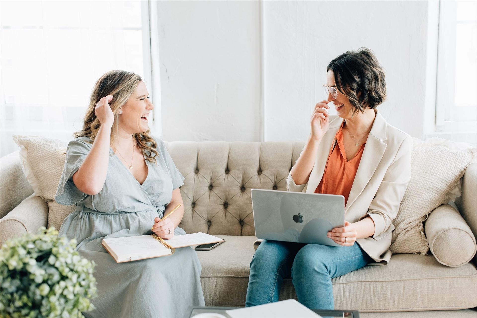 Two women are sitting on a couch with laptops and talking to each other.