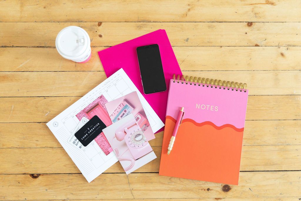 Pink and orange notebook with pen, phone, pink folder, and photos on a wood surface.