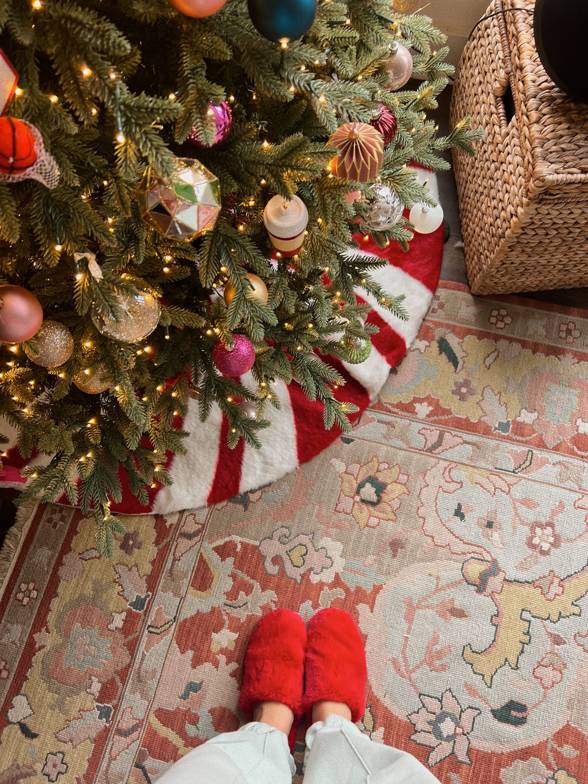 Feet in red slippers standing before a decorated Christmas tree with a red and white tree skirt on a patterned rug.