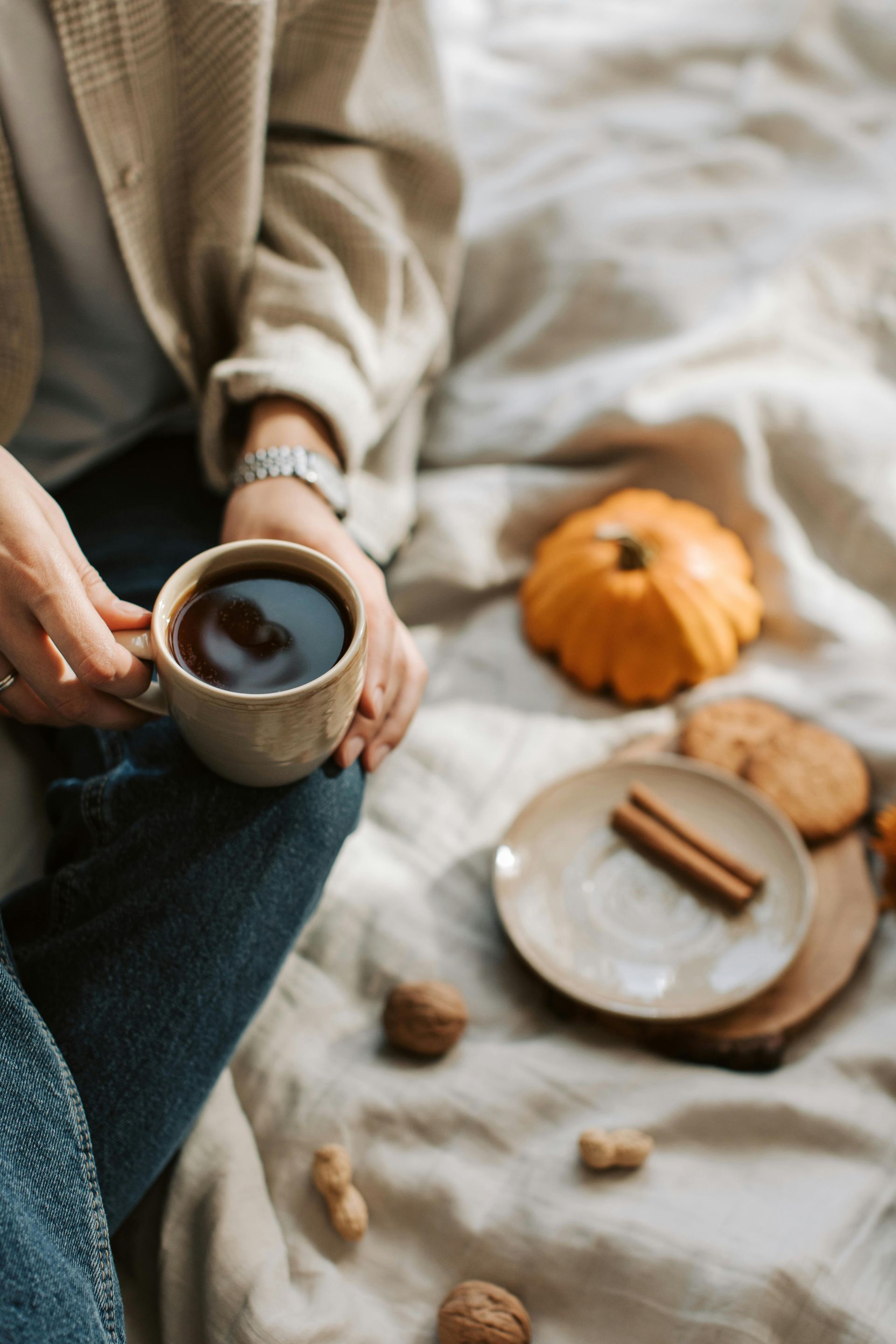 Person holding coffee cup, seated with pumpkin, cookies, and cinnamon on bed.