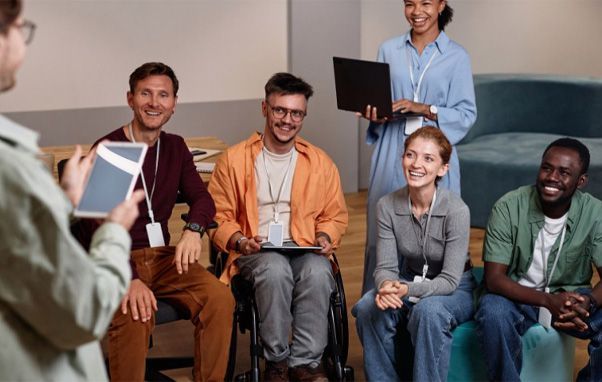 Group of people in a circle, one in a wheelchair, smiling during a meeting. A person holds a tablet.