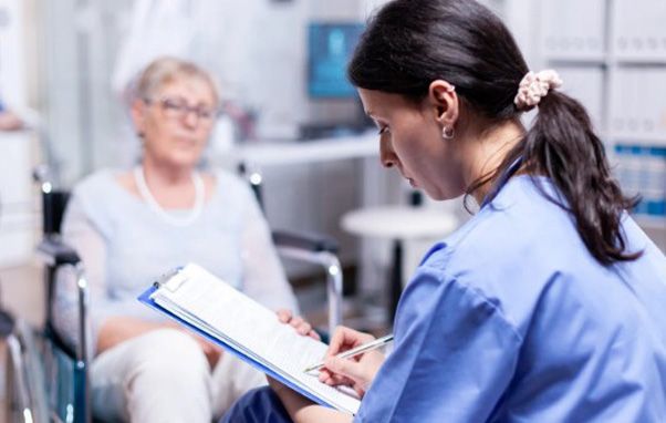 Nurse in blue scrubs writing on a clipboard while talking to a woman in a wheelchair indoors.