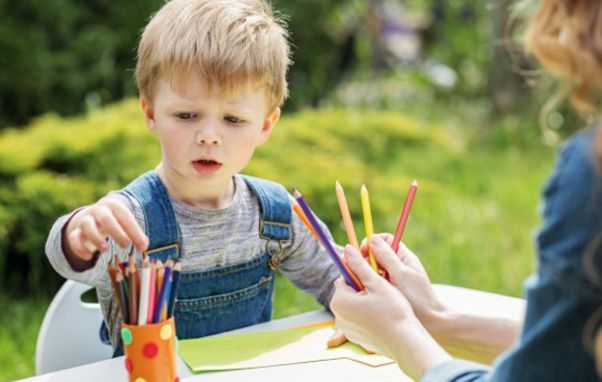 Boy in overalls selecting a pencil from a hand, outdoors, next to a craft table with crayons.