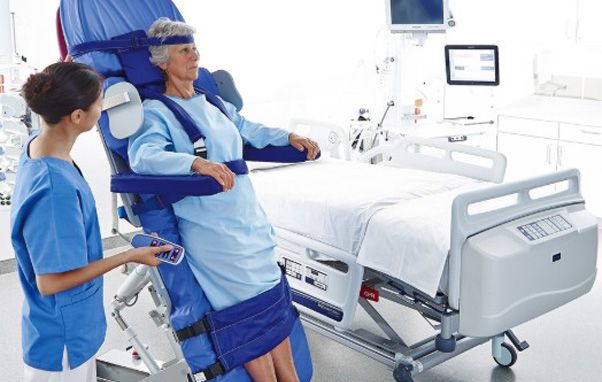 Woman in medical chair, nurse adjusting controls. Hospital setting with bed, equipment. Blue tones, serious expressions.