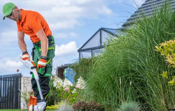 Man in orange shirt and green cap using a trimmer on tall grass in a garden setting.