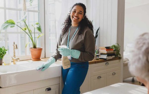 Woman in blue gloves smiles while cleaning a kitchen sink near a sunny window.