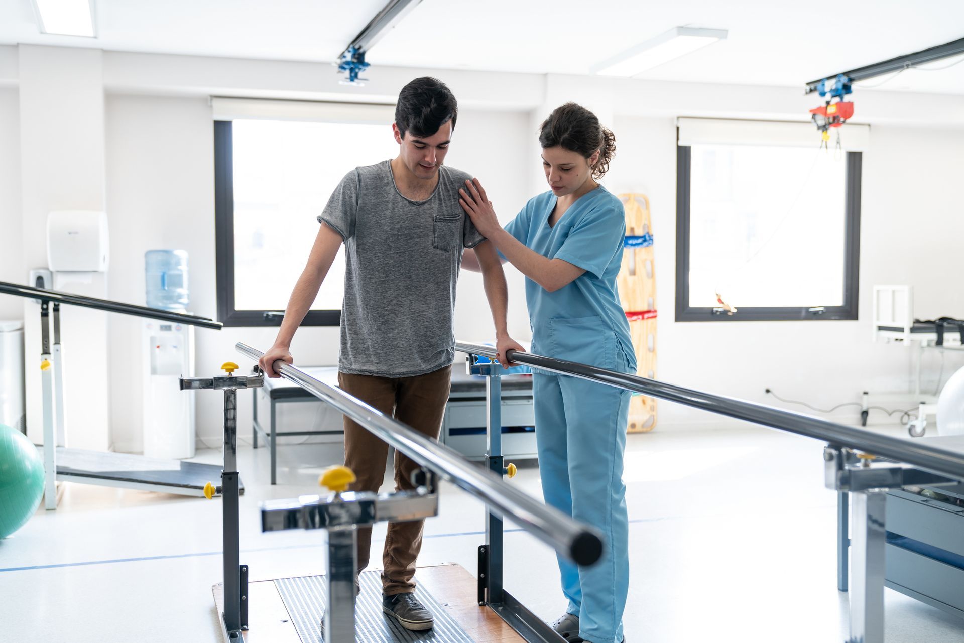 A nurse is helping a young man walk on parallel bars in a hospital.