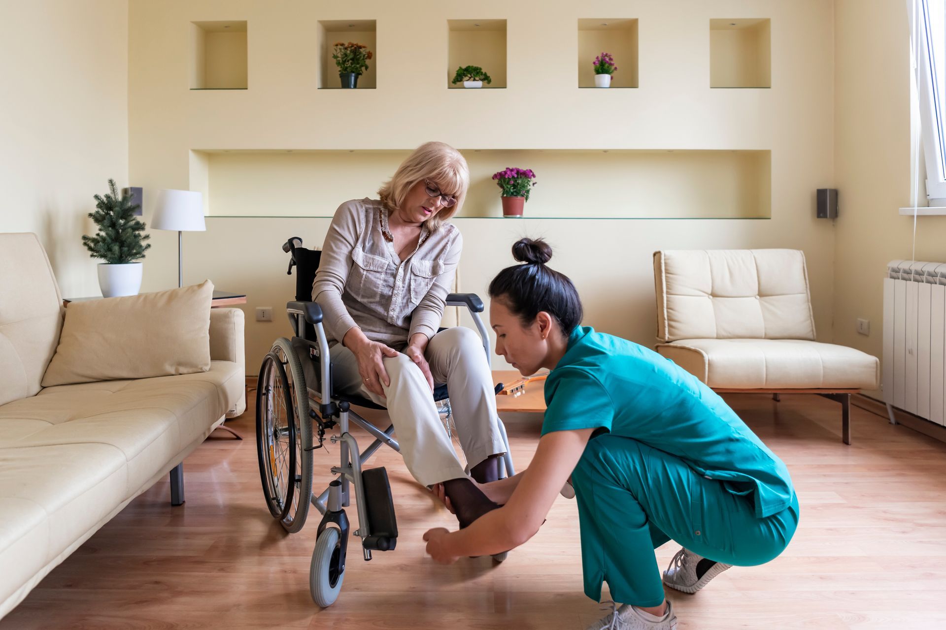 A caregiver in a blue uniform is helping a woman in a wheelchair to put shoes on.