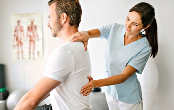 Woman in blue scrubs providing physical therapy to a man, touching his back and shoulder. Light setting.