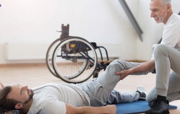 Man receiving physical therapy; therapist assisting with leg stretch. Wheelchair in the background.