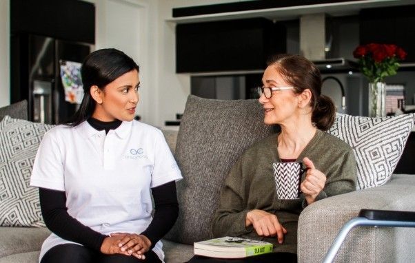 Caregiver and senior woman on a sofa, talking. The woman holds a mug and has a book.