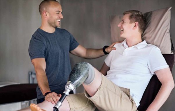 A man with a prosthetic leg smiles as another man puts a hand on his shoulder in a medical setting.