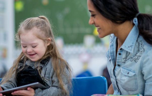 Girl with Down syndrome smiles at a tablet, teacher looks on, classroom setting.