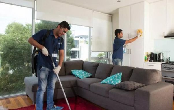 Two people cleaning a living room. One vacuums the floor, the other wipes a cabinet.