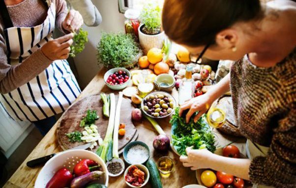 Two women preparing food in a bright kitchen with vegetables and fruits on a wooden countertop.