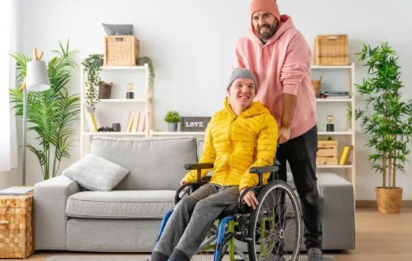 Man pushing a wheelchair with person inside, both smiling. Indoors, living room setting.