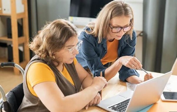 Woman in wheelchair works on laptop with assistance from another woman pointing at the screen.