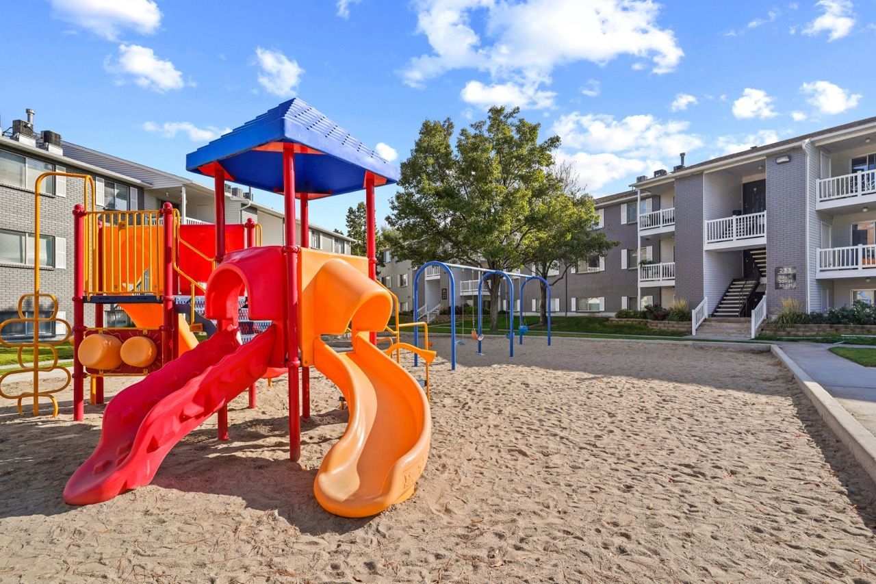 Colorful playground with slides and climbing structures on sand, with apartment buildings in the background.